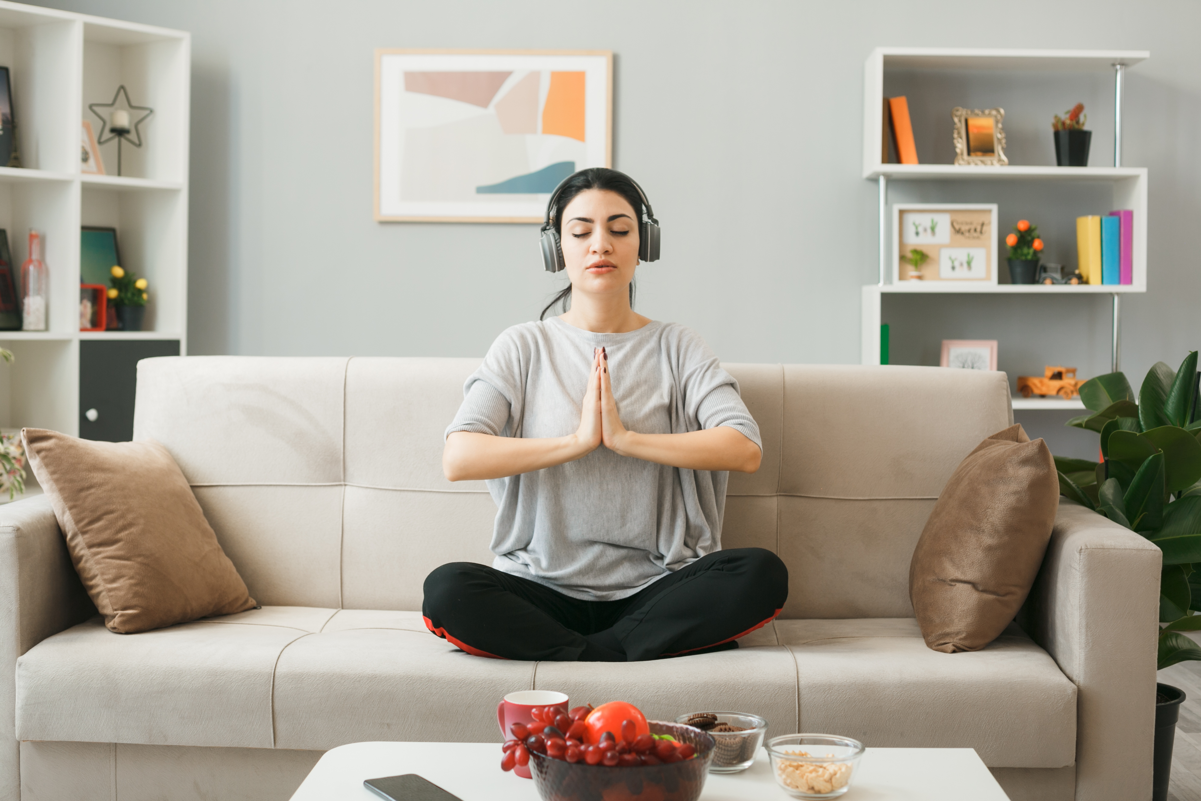 confident young girl wearing headphones doing yoga sitting on sofa behind coffee table in living room